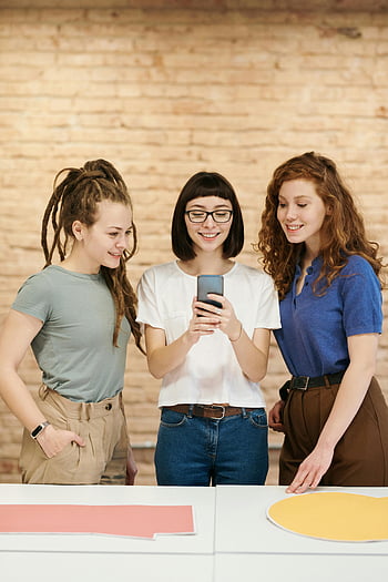 スマートフォンを探している女性、 3人の若い女性、 スタイリッシュなルックス、 幸せな瞬間、 室内でのスマートフォン、 レンガの壁の背景、 カジュアルな服装、 グループの相互作用、 現代の技術、 屋内設定、 笑顔の女性