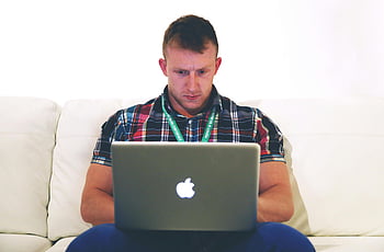 man using Macbook, white sofa, young man, typing on laptop, seated on couch, indoors, focused, plaid shirt, Apple laptop, casual setting, comfortable environment