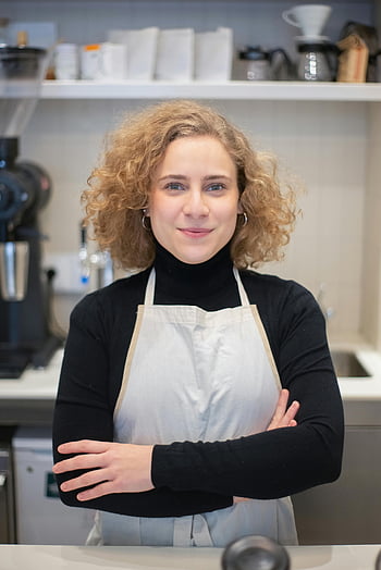 woman wearing apron, cheerful barista, stylish coffee shop, folded arms, confident pose, coffee shop worker, apron at work, barista at work, coffee shop interior, professional barista, smiling barista