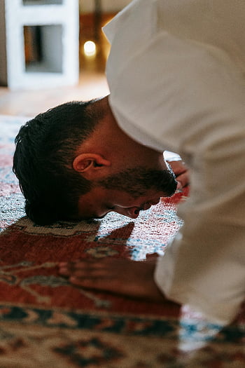 man, white thobe, bowing, prayer, red and blue rug, traditional attire, patterned rug, indoor setting, cultural practice, religious act, spiritual moment