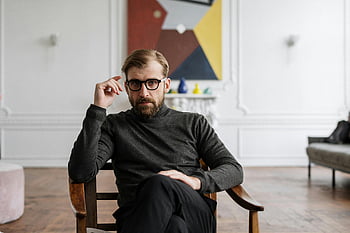 man in zwarte trui, bruine houten stoel, modern interieur, abstracte kunst, man met een bril, zittende man, eigentijdse kamer, Stijlvolle man, binnenomgeving, Casual pose, artistieke achtergrond