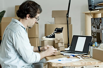 man preparing packages, businessman packing products, managing online store, desk with laptop, packaging products, online store management, small business owner, product packaging, e-commerce business, home office setup, shipping products