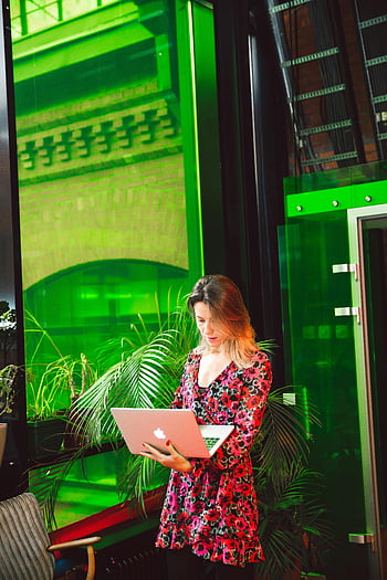 woman in floral dress, using Macbook, working remotely, laptop, vibrant green backdrop, young woman, indoor setting, remote work, floral dress, green background, laptop user