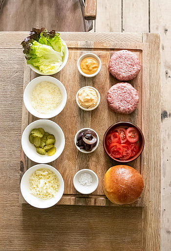 burger ingredients, wooden table, wooden chopping board, hamburger patties, lettuce, cheese, pickles, tomatoes, onions, mayonnaise, salt