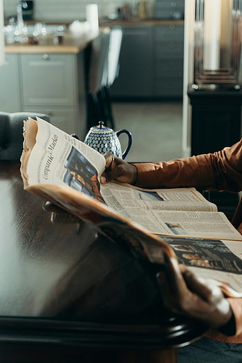 persona che legge il giornale, tavolo di legno, impostazione interna, teiera, Lettura per adulti, giornale, sfondo della cucina, Atmosfera accogliente, routine quotidiana, Scena casalinga, abitudine alla lettura