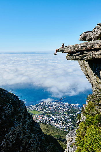 personne assise sur une falaise, falaise de montagne, vue sur l'océan, ville ci-dessous, une vue à couper le souffle, bord de la falaise, paysage pittoresque, aventure en plein air, photographie de nature, photographie de voyage, vue panoramique