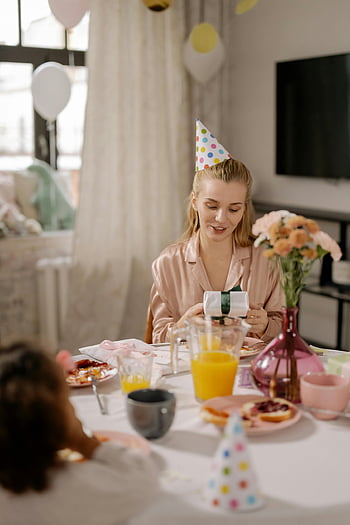 mujer, mangas largas rosa, sombrero de fiesta, sosteniendo un regalo, desayuno, celebración de cumpleaños, globos, mesa festiva, celebración matutina, apertura de regalos, ambiente festivo