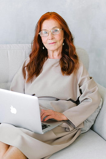 elderly woman, red hair, using laptop, sitting on sofa, indoors, charming, glasses, casual outfit, silver laptop, comfortable setting, home environment