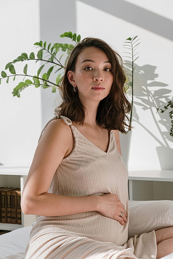 zwangere vrouw, beige nachthemd, zittend op bed, hand op de buik, Gezellige slaapkamer, Moeder in verwachting, zwangerschapsmode, zwangerschapsstijl, binnenomgeving, natuurlijk licht, plant decor