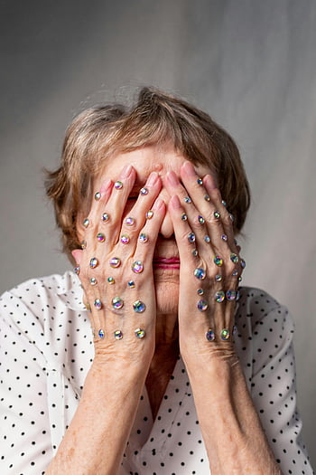 oudere vrouw, wit overhemd, bedekkend gezicht, handen, sprankelende strass, polka stippen, emotionele expressie, grijze achtergrond, portret, detailopname, senior