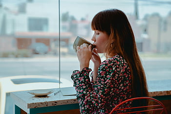 コーヒーを飲みながら女性、 スタイリッシュなカフェ、 朝の雰囲気、 平穏な瞬間、 コーヒーカップ、 花柄ドレス、 カフェ設定、 屋内シーン、 自然光、 都市の背景、 赤い椅子
