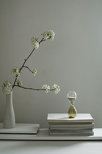 Still Life minimalista, ramo floreale, clessidra, libri impilati, vaso bianco, sfondo bianco, arredamento moderno, composizione semplice, estetica pulita, fotografia al coperto, luci soffuse