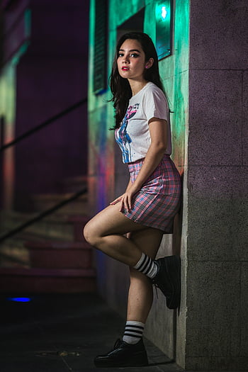 moderigtig ung kvinde, læner sig på væggen, by stil, plaid nederdel, grafisk t-shirt, Stilfuldt outfit, nat scene, moderne mode, gadestil, afslappet look, trendy tøj