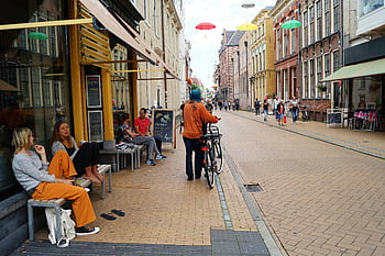 scena di strada, persona che tiene una bicicletta, costruzione, gente che cammina, bike, negozi vivaci, caffè, giorno soleggiato, ombrelli di strada, vita di città, posti a sedere all'aperto