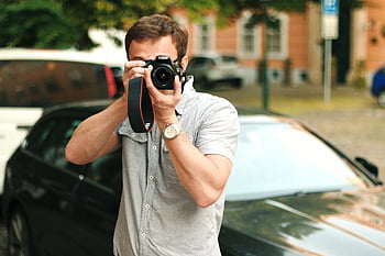 man taking picture, DSLR camera, city setting, outdoors, beside car, photographing, urban photography, street photography, male photographer, casual attire, daytime scene
