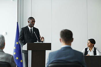 man in black suit, speaking, formal business meeting, diverse speakers, conference room, podium, audience, professional event, corporate presentation, European Union flag, business conference