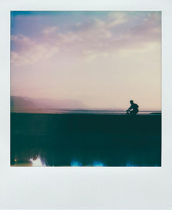 Polaroid picture, person biking, white clouds, vintage-style silhouette, cyclist, twilight, serene sky, coastline, beach scene, outdoor activity, evening ride