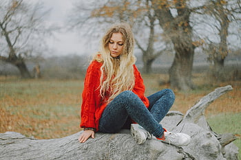 žena v červeném svetru, sedí na protokolu, blond vlasy, venkovní scénu, džíny, tenisky, přírodní pozadí, Nastavení podzimu, Neformální outfit, uvolněná pozice, kmen stromu