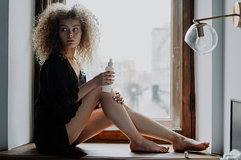 Mulher de vestido preto, sentado na janela, janela de madeira marrom, jovem mulher, cabelo cacheado, segurando um frasco de loção, cena interna, luz natural, pose casual, configuração relaxada