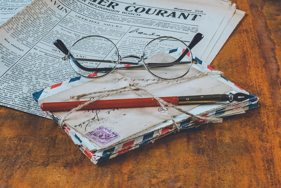 historical letters, fountain pen, vintage wooden table, eyeglasses, newspaper, antique desk, old correspondence, writing tools, vintage eyewear, historical artifacts, classic desk setup
