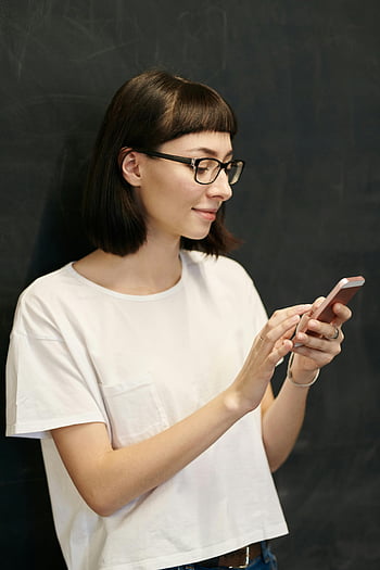 γυναίκα που κατέχει smartphone, νέα γυναίκα, Γυαλιά, χρησιμοποιώντας smartphone, χαμόγελο, Φόντο μαυροπίνακα, Casual ντύσιμο, λευκό μπλουζάκι, κοντά μαλλιά, εσωτερική ρύθμιση, χρήση τεχνολογίας