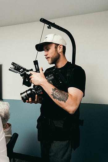 muž v černém, tričko posádky krku, Držení černé kamery DSLR, Vnitřní video natáčení, Provozní vybavení kamery, profesionální fotograf, video produkce, kameraman, fotografování v interiéru, černý fotoaparát, Nastavení videokamery