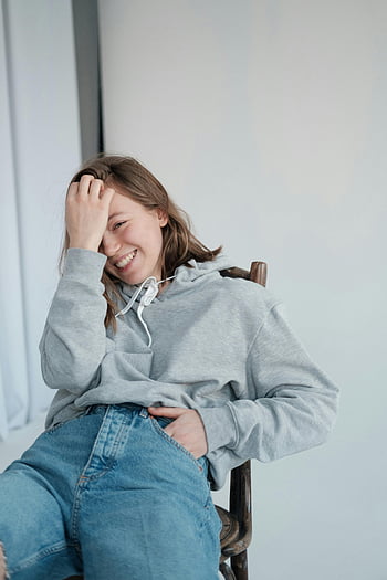 radostná žena, sedí na židli, ruku v kapse, Mladá žena, šedá mikina s kapucí, usmívající se, Nastavení studia, pozitivita, radost, příležitostná pozice, džíny denim