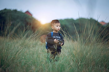 shallow focus photography, boy, green grass, holding camera, sunset, toy camera, backpack, grassy field, young boy, outdoor scene, nature photography