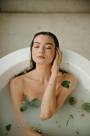 Frau in der Badewanne, Entspannende Selbstpflege, Gelassene Frau, Blütenblätter, Wasserbad, weiße Badewanne, Selbstpflegemoment, friedliches Bad, Frau, die Bad genießt, Bad mit Blütenblättern, Entspannungsbad