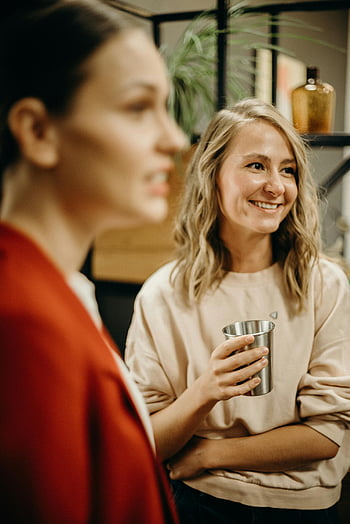 due donne, raduno indoor, conversazione casuale, donne sorridenti, tenendo bevande, impostazione interna, abbigliamento casual, Interazione sociale, Atmosfera amichevole, Evento indoor, bevande casuali