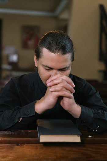man praying, church pew, black clothing, hands interlocked, holy book, religious setting, prayer, spiritual moment, indoor church, faith practice, contemplation