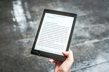 e-book reader, black e-reader, person holding, wet pavement, text display, outdoor reading, digital book, e-reader device, handheld device, reading technology, e-reader screen