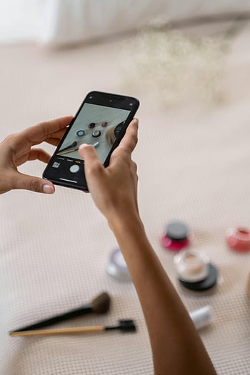 pessoa tirando foto, smartphone preto, produtos de maquiagem, cama, captura de imagem, produtos de beleza, itens cosméticos, pincéis de maquiagem, cuidado pessoal, fotografia de estilo de vida, fotografia de produto