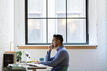 man, grijs shirt met lange mouwen, gefocust, mannelijke professional, werkend, laptop, bureau, groot raam, binnenwerkruimte, professionele setting, concentratie