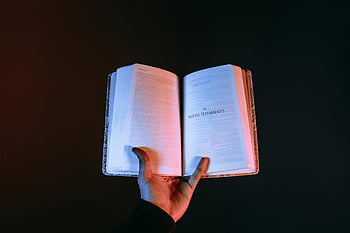hand holding Bible, open Bible, New Testament, dark background, religious book, spiritual text, holy scripture, reading Bible, faith symbol, religious image, spiritual reading