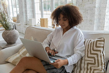 Afroamerikanerfrau, Lockige haare, Tippen auf Laptop, Gemütliches Sofa, Helles Wohnzimmer, Beruhige junge ethnische Dame, Gemütliche Wohnung, konzentriert sich auf den Laptop, Ethnische Dame, gemütliches Wohnzimmer, Helle Wohnung