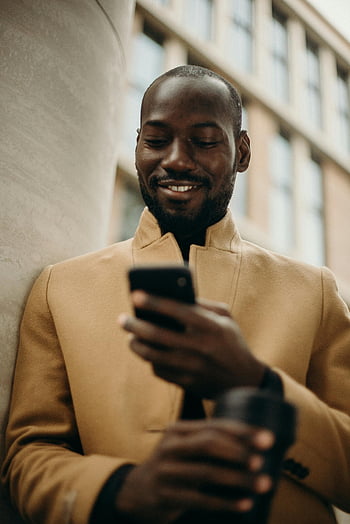 uomo sorridente, smartphone, cappotto elegante, caffè, pilastro di cemento, allegro, all'aperto, contesto urbano, uomo moderno, Uso tecnologico, momento casuale