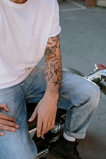 man in white shirt, denim jeans, arm tattoos, seated on motorcycle, casual style, outdoor setting, urban scene, motorcycle rider, tattooed arm, casual outfit, street style