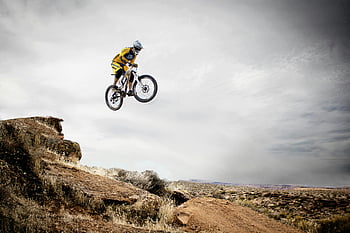 cyclist, daring jump, rocky cliff, cloudy sky, mountain biking, extreme sports, outdoor adventure, aerial stunt, bike jump, adventure photography, nature backdrop