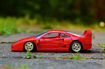 Red Ferrari F40, die-cast toy, miniature toy car, sidewalk, greenery backdrop, detailed model, red sports car, toy vehicle, outdoor setting, classic car toy, Ferrari F40 Coupe