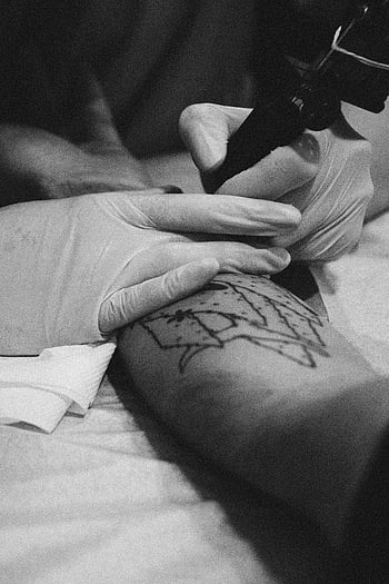 grayscale photography, tattoo artist, tattoo machine, black and white, close-up photo, arm tattoo, studio setting, tattooing process, gloves, artistic work, monochrome image