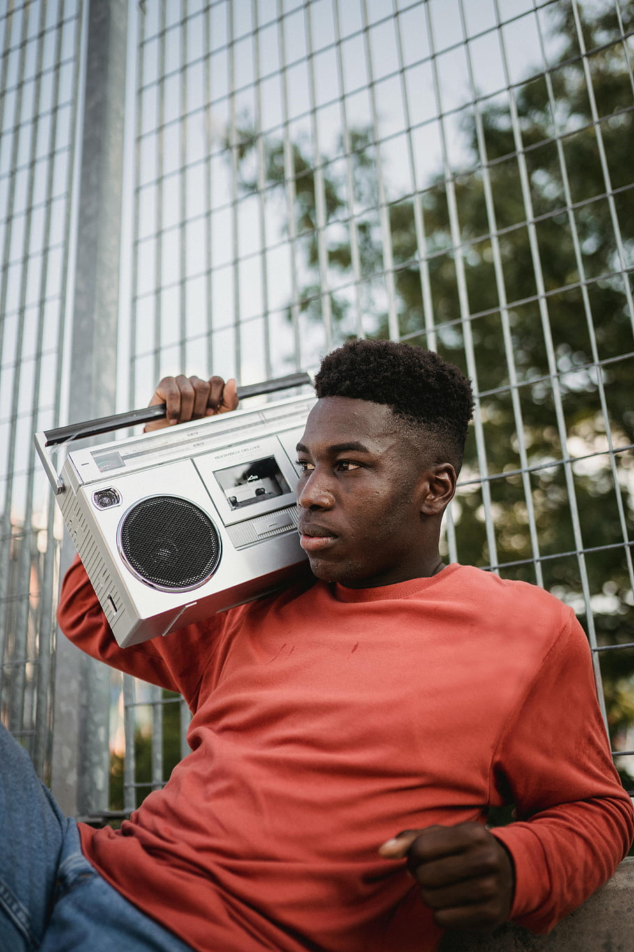Černoch, Vintage boombox, červený svetr, venkovní nastavení, kovový plot, Poslech rádia, mladý muž, Trskový výraz, Retro zvukové vybavení, městské scény, Nostalgická vibrace