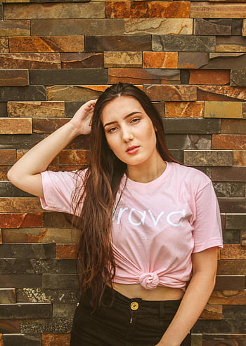 woman in pink shirt, confident pose, brick wall background, fashionable look, young woman portrait, pink crew-neck shirt, stone wall, stylish outfit, casual fashion, confident woman, trendy style