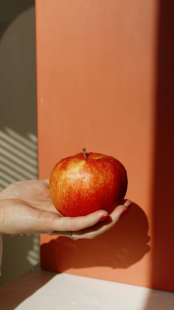 červené jablko, ruka drží jablko, oranžové pozadí, detailní fotografie, přirozené světlo, stíny, ovocné fotografie, Zdravé stravování, stálý život, zářivé barvy, Minimalistický styl