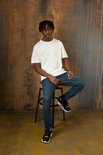 young black man, sitting on chair, wooden wall, looking at camera, confident young man, stylish outfit, modern fashion, relaxed pose, denim jeans, white t-shirt, black sneakers