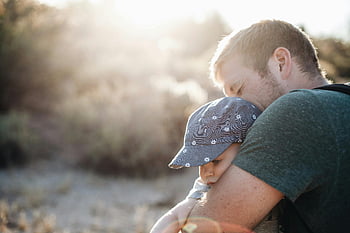 Otec objímá dítě, slunečné odpoledne, venkovní fotografie, Den otců, milující objetí, dítě na sobě čepici, Rodinný okamžik, přirozené světlo, Emocionální spojení, rodičovství, venkovní nastavení