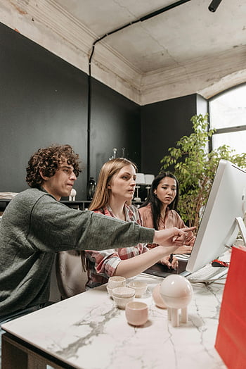 man in gray sweater, two women, modern office, diverse team, strategy discussion, technology use, creativity enhancement, office setting, collaborative work, professional meeting, computer screen
