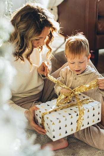 ibu dan anak laki-laki, Hadiah Pembuka, hadiah Natal, semangat liburan, momen yang menyenangkan, Adegan dalam ruangan, ikatan Keluarga, Suasana yang meriah, pembungkus kado, pita emas, kotak hadiah putih