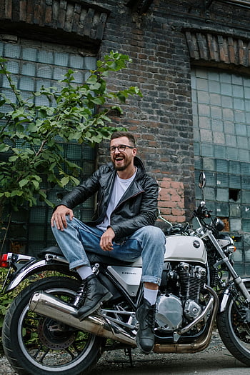 smiling man, leather jacket, motorcycle, brick wall, outdoor, casual style, glasses, urban setting, bike, rider, streetwear