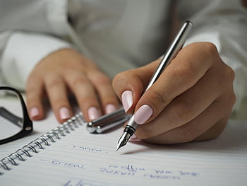 woman writing, white long sleeved shirt, holding pen, writing on paper, notebook, fountain pen, pink nails, close-up, hands, glasses, desk